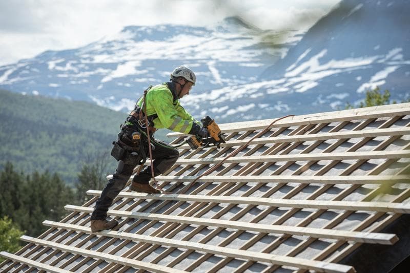 Trabalhador usa uma pregadora DEWALT em telhado inclinado ao ar livre, equipado de segurança, montanhas ao fundo.