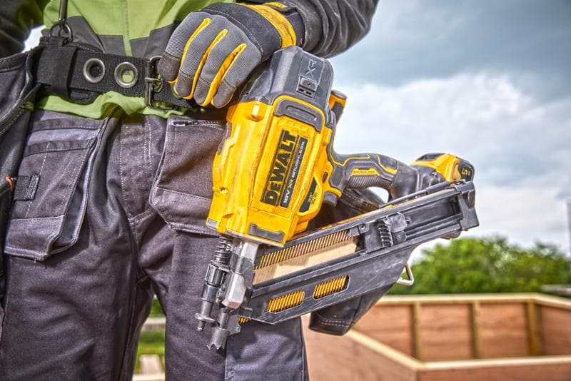 Pessoa segurando uma clavadora DEWALT DCN930N, com luvas e vestimenta de trabalho em um canteiro externo.