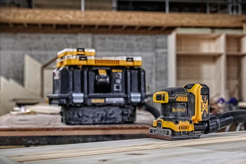 Lixadeira sem fio DEWALT em madeira, caixa de armazenamento ao fundo em oficina.
