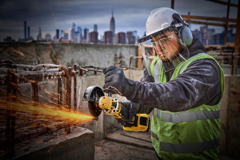 Trabalhador usa esmerilhadeira DEWALT DCG405P2 na obra, com equipamento de segurança, cidade ao fundo.