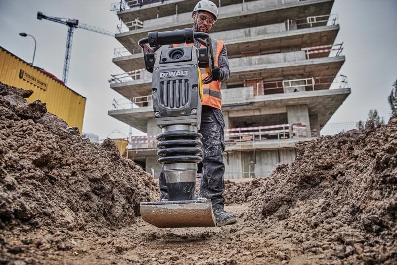 Compactador DEWALT em obra, operado por trabalhador com equipamento de segurança, rodeado de terra e prédios.