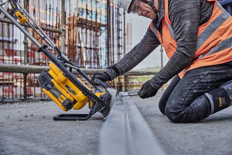 Trabalhador usa ferramenta amarela DEWALT para cortar metal na obra, veste colete laranja e equipamento de segurança.