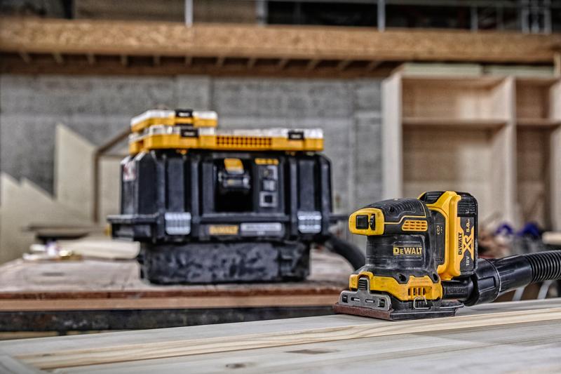 Lixadeira sem fio DEWALT em madeira, caixa de armazenamento ao fundo em oficina.