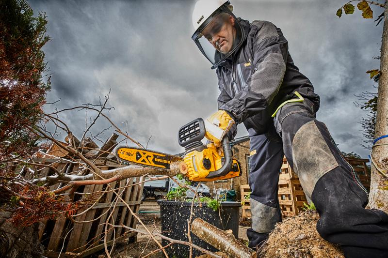 Pessoa equipada com proteção usa motosserra DEWALT para cortar galhos ao ar livre em dia nublado.