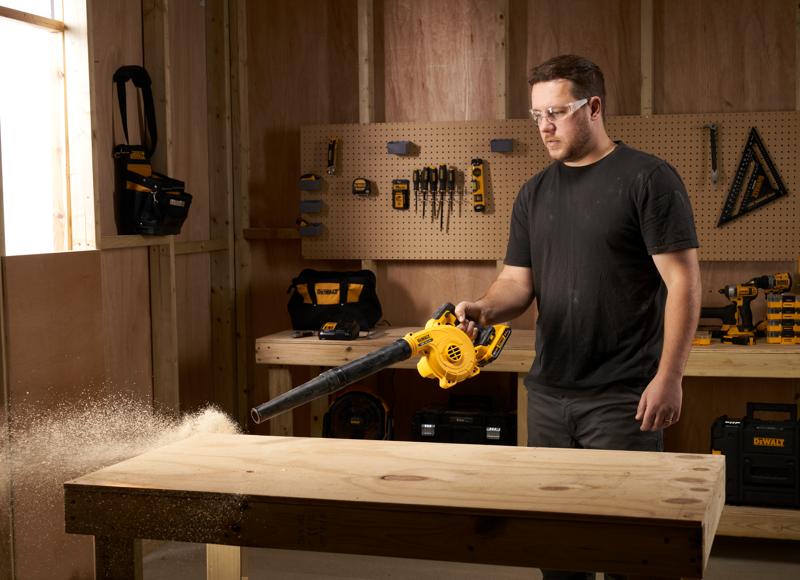 Pessoa usa soprador DEWALT DCV100 para remover serragem de uma bancada de madeira em oficina.