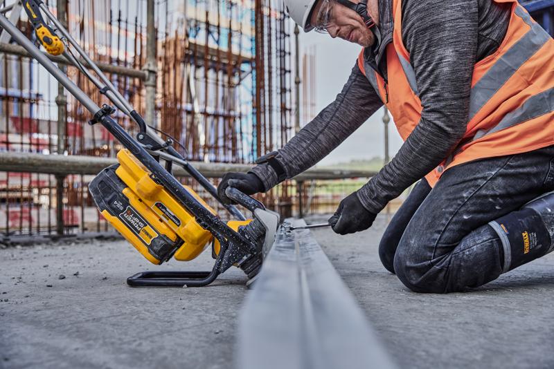 Trabalhador usa ferramenta amarela DEWALT para cortar metal na obra, veste colete laranja e equipamento de segurança.